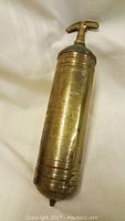 Photo of brass mini fire extinguisher showing engraved instructions on the cylindrical body with T-handle top and ridged bottom.