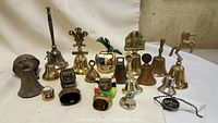 Wide shot of entire collection of 14 vintage hand bells of various materials and sizes, including brass, metal, and leather accessories on a white fabric background.