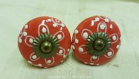 Pair of bright orange round porcelain knobs with white hand-painted floral design and metal mounting hardware, placed on flat surface.