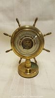Front view of brass barometer shaped like a ship's wheel with six handles, mounted on a pedestal base showing the dial and central decorative image.