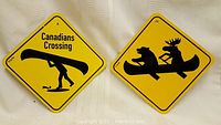 Two yellow Canadian themed tin signs on a white fabric background. Left sign shows a person carrying a canoe and the text 'Canadians Crossing'. Right sign shows a bear and moose in a canoe.