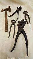 Five antique hand tools on white fabric: hammer with wooden handle and multi-prong toothed head, adjustable black pliers, two small tongs/pincers with ring handles, vintage wire cutter or crimper tool.