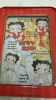 Close-up of the front of the rectangular red tin handheld game showing colorful Betty Boop artwork with angel and devil motifs and text phrases.