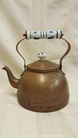 Front view of the old copper kettle showing the curving spout, hinged handle with Delft-style ceramic grip, and ceramic lid knob with blue floral design.