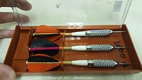 Top view of three vintage darts laid inside orange base plastic case showing metal barrels, white shafts, and black-orange and black-pink flights.