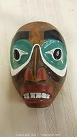 Mini native styled hand-carved mask with painted details featuring expressive eyes and grimacing mouth on natural wood surface.