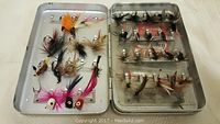 Open metal box containing assorted fly fishing flies in bright colors and feathered patterns on both sides.