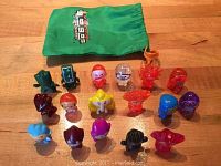 Top-down photo showing 16 colorful Evolution Gogos Crazy Bones figurines arranged on a wood surface, next to a green drawstring storage bag with printed logo.