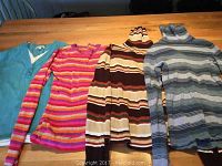 Seven ladies sweaters laid out on a wooden table, showing various colors, stripes, and styles such as turtlenecks and v-necks.