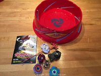 Wide photo showing the red Beyblade arena, four Beyblades, two launchers, one red ripcord, and one manual.