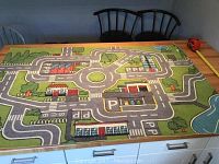 Photo of rectangular children's play rug with road, parking, and buildings design on table with chairs and tape measure nearby.