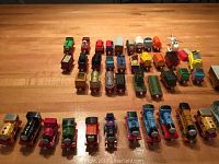 Overview of arranged Thomas & Friends toy engines and various cars laid out on wooden surface