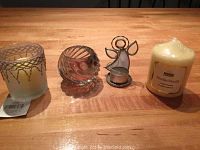 Front view of the four items on a wood table showing their shapes and sizes: frosted glass candle holder with yellow candle, clear glass swirled candle holder with red candle, metal angel figurine tealight holder with tealight, large vanilla-scented candle labeled Reglal