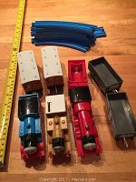 Photo showing the three Thomas engine trains in blue, gold and red, two beige train cars, two black train cars, a red station piece, and six curved blue plastic track pieces with a measuring tape beside it for scale