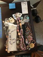 Overhead photo of assorted costume jewelry necklaces on a wooden table, including pink, peach, black, and multicolor beads with tags visible.