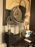 Photo showing the acrylic clock with curved clear tubing mounted on a dark textured base, and a nude female torso statue in the background.