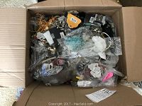 Top view of box filled with assorted jewelry items including necklaces, earrings, beads, and bangles in plastic packaging.