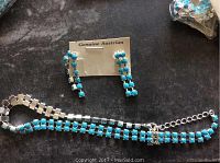 Two blue crystal dangling earrings with genuine Austrian crystals on a card, placed beside a blue crystal necklace with silver-tone links on black fabric background.