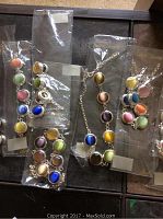 Photo of multiple multi-colored jewelry pieces - necklaces and bracelets, each with round colorful domed beads in silver-tone metal settings, all new in clear plastic packaging, displayed on dark surface.