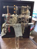 Clear acrylic T-shaped display stand filled with multiple necklaces and bracelets, each tagged new, showing variety of styles and colors including metallic chains and beaded designs.
