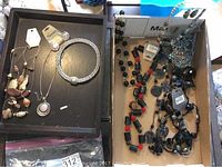 Mixed collection of necklaces and a mesh bracelet on a black tray and a cardboard box. Many have tags attached, showing a mix of styles and materials in new condition.