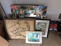 Photo showing multiple framed art pieces and a decorative mirror leaning against the wall: Cape Cod map, floral art, cartoon print, Life magazine themed mirror.