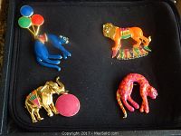 Four vintage enamel animal pins laid out on black fabric, showing a blue cat with balloons, an orange lion, a gold-toned elephant, and a pink spotted cat.