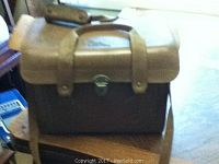 Leather camera carrier with handle sitting on a wooden chair, showing its rough texture and buckle clasp closure.