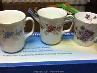 Photo showing three china mugs with floral and violet patterns on a box for a paper shredder, featuring delicate handles and fine china material.