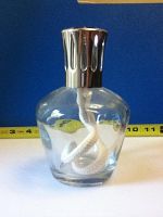 Full view of translucent glass Lampe Berger fragrance lamp with chrome cap and white coiled wick inside, against blue background with a measuring tape behind showing approximate height.