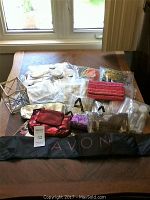 Wide shot of Avon miscellaneous products laid out on a wooden table showing furry white hot water bottles, mesh vegetable bags, several clear bathroom organizers, fabric wallets, and other small items including decorative and fabric bags.