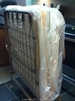 Folded single bed on metal frame with mattress visible through plastic protective cover, positioned vertically on wheels.