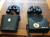 Photo showing both sets of binoculars with their respective cases on a wood surface; Bushnell set on right with black hard case, Carl Wetzlar set on left with soft greenish case.