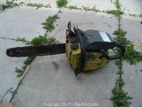 Full view of the Pioneer chainsaw lying on concrete ground with vegetation around. Shows yellow body and chain bar length.