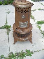 Full view of the rusty, dented vintage oil burning stove standing on an outdoor tile surface with grass nearby.