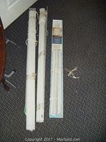 Two rolled white Altex heavy pull down blinds laid side by side on a carpeted floor alongside a packaged Mainstays mini blind with pull cords visible, showing size and texture detail.