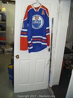Front view of the Edmonton Oilers blue, orange, and white hockey jersey hanging on a door, showing full front and sleeves with team logo and alternate captain patch.