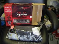Photo showing the original red and black Sony Xplod box with model CDX-MP40, along with the wiring harness and installation kit in plastic bags on a chair.