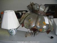 Photo showing hanging lamp with floral glass panels and metal edging, and the table lamp with floral ceramic base and pleated white shade placed on a table