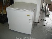 Full view of the white Salton mini fridge on floor showing front, side, and top edges with power cord visible.