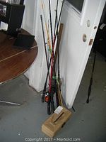 Image showing four fishing poles standing against a door, with visible reels on three poles (open face) and one closed face reel with non-matching pole end. An extra pole is partly visible. A beige Old Pal tackle box is on the floor in front of the poles.