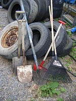 Wide angle view showing rakes, shovels, and edger leaning on tires outdoors on gravel
