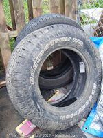 Photo showing BF Goodrich Rugged Trail T/A tire with white lettering and tread visible, leaning against a fence with multiple tires behind it.