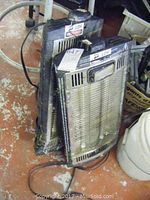 Two electric ceiling mount quartz heaters stacked on the floor, showing metal grille fronts, black sides, wires and cords on the ground, visible wear and dents.
