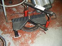Diagonal view of the Hempe Manufacturing metal mitre saw jig showing ribbed angled platforms, red clamp handles, and semi-circular angle gauge.