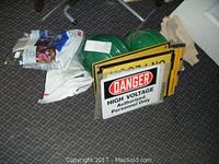 Photo showing assorted safety signs, including a 'Danger High Voltage Authorized Personnel Only' sign, stacked together on carpeted floor with two green hard hats.
