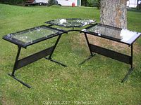 Wide view of metal frame desk pieces arranged on grass, showing two rectangular sections and a corner section with clear glass tops and black metal frames.