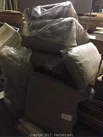 Four outdoor patio furniture cushions stacked and individually wrapped in plastic, dark color, square shape.