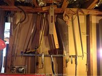 Photo showing 12 assorted hand tools hanging on a wooden wall including saws, crowbar, cable cutter, axe, hammer, wrench, clamps, level, and a hacksaw frame.