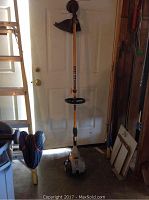Ryobi gas-powered weed eater standing upright in a garage against a white door, showing full length and handle.
